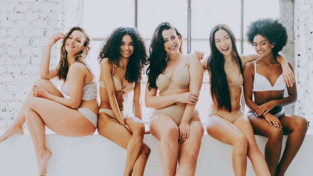 Five young women of different ethnicities and body types smiling confidently in natural-toned underwear, celebrating real, healthy skin.